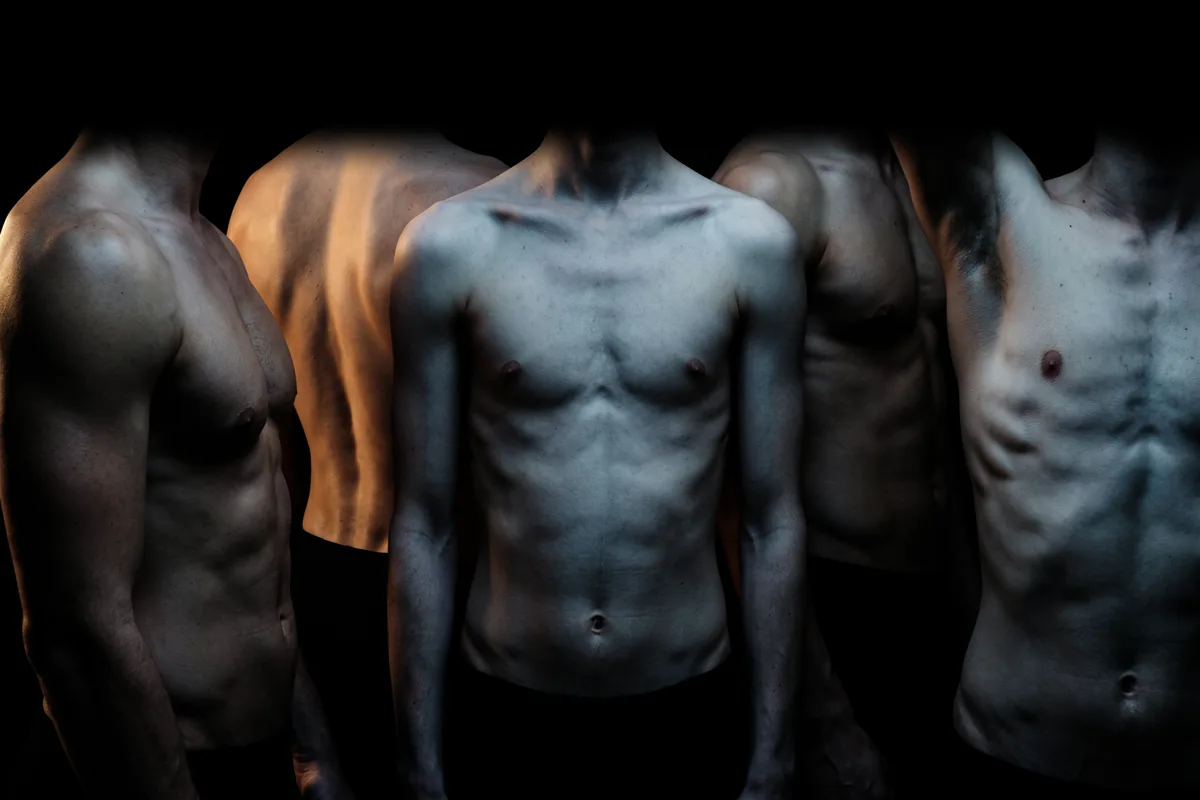 Five bare-chested men standing close together in the dark, seen from the shoulders down: a slim man faces forward between broader, more muscular men.